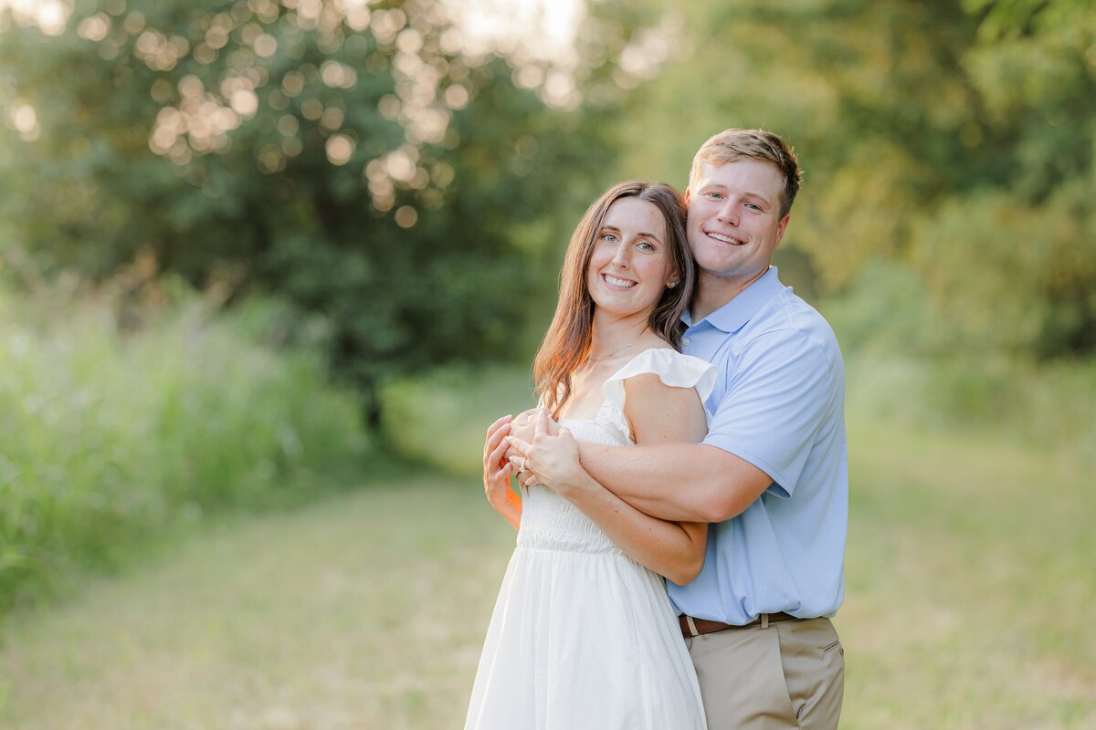 A man in a blue polo hugs his fiancee from behind in a white dress at sunset