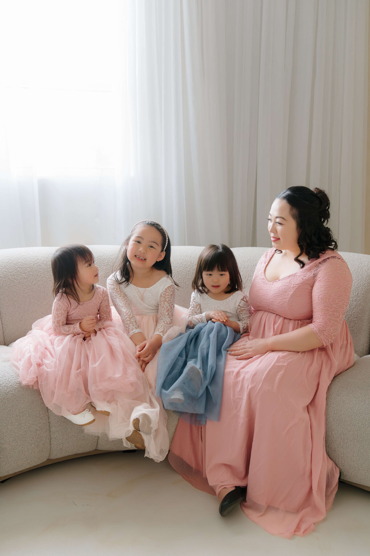 A woman and three children in elegant pink dresses, gracefully seated together on a plush couch, exuding warmth and joy.