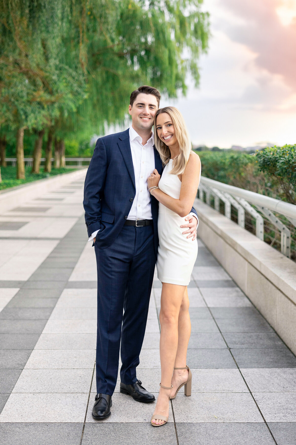 A couple standing on a tiled walkway surrounded by greenery. The man is wearing a dark suit, and the woman is in a white dress and heels. They smile warmly at the camera with trees in the background and a cloudy sky.
