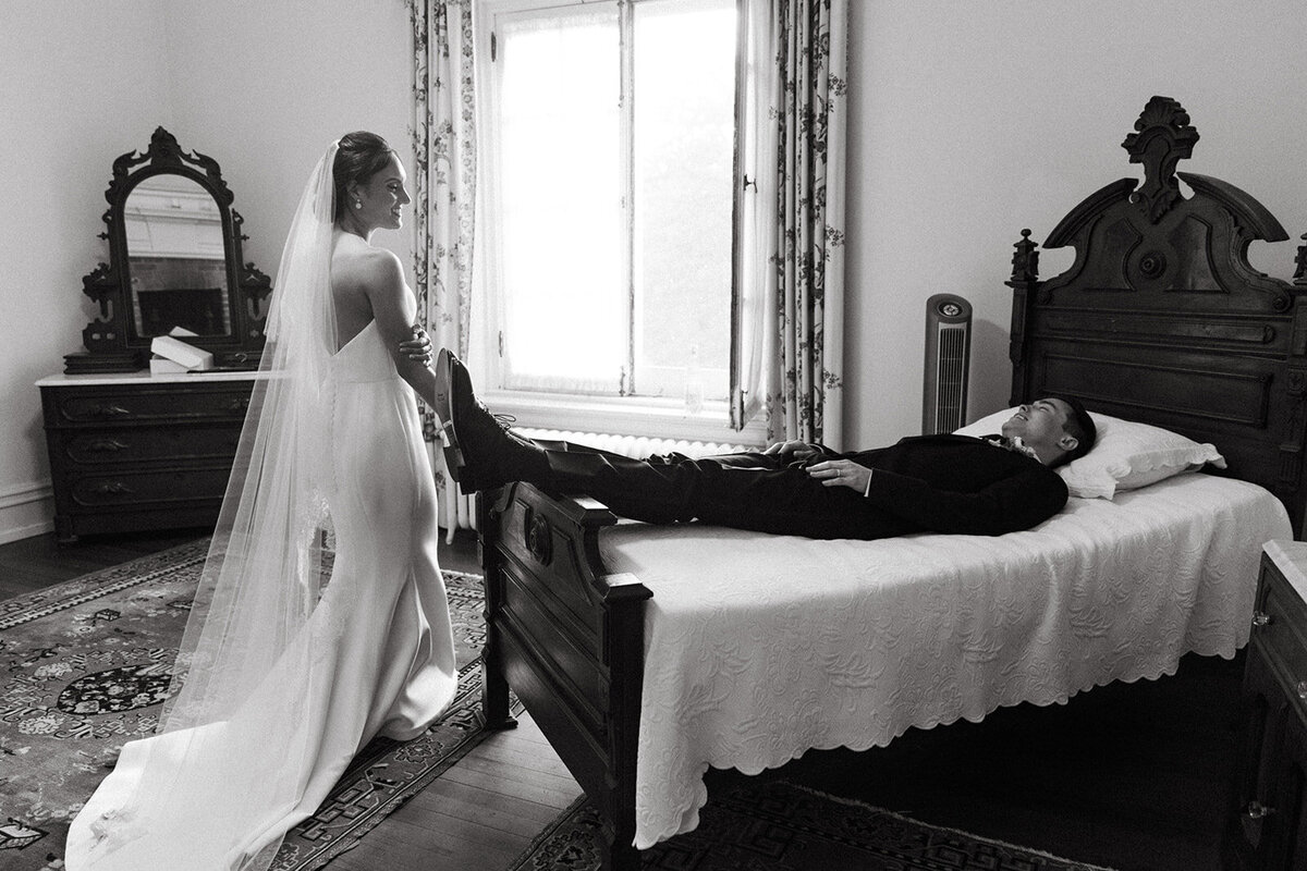 Bride in a white dress and veil holding groom's foot on a bed