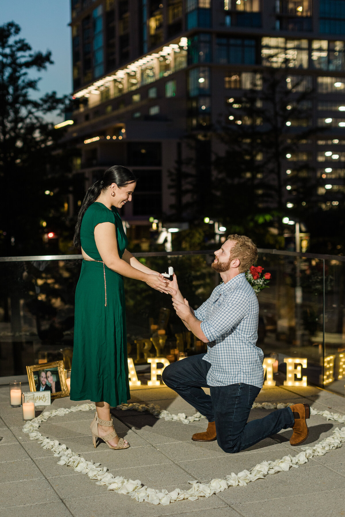 A photo of a man proposing to his future fiancé. The man is down on one knee with the engagement ring, and the couple is surrounded by a heart shaped ring of flower pedals as well as photos, candles, and light up letters that say "Marry Me" in the background. The woman on the left is wearing a green dress and high heels while the man on the right is wearing a blue and white checkered dress shirt and jeans.