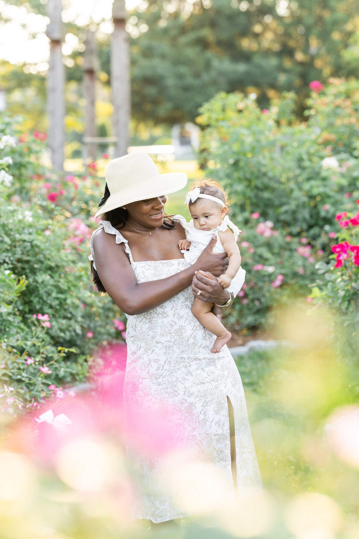A mother and her baby share a tender moment in a vibrant green garden filled with blooming flowers. The mother is smiling at her baby as she gazes down at the flowers with curiosity. 