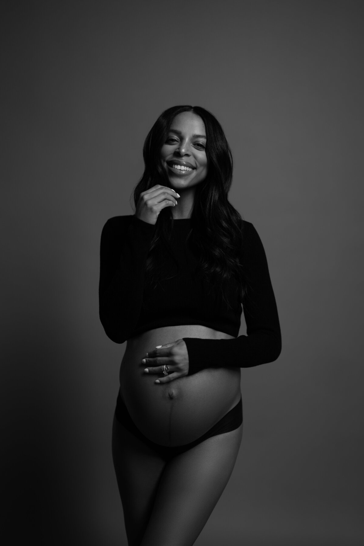 A maternity portrait featuring the mother-to-be in a fitted black outfit, holding her bump with a gentle smile, taken in a NYC studio.