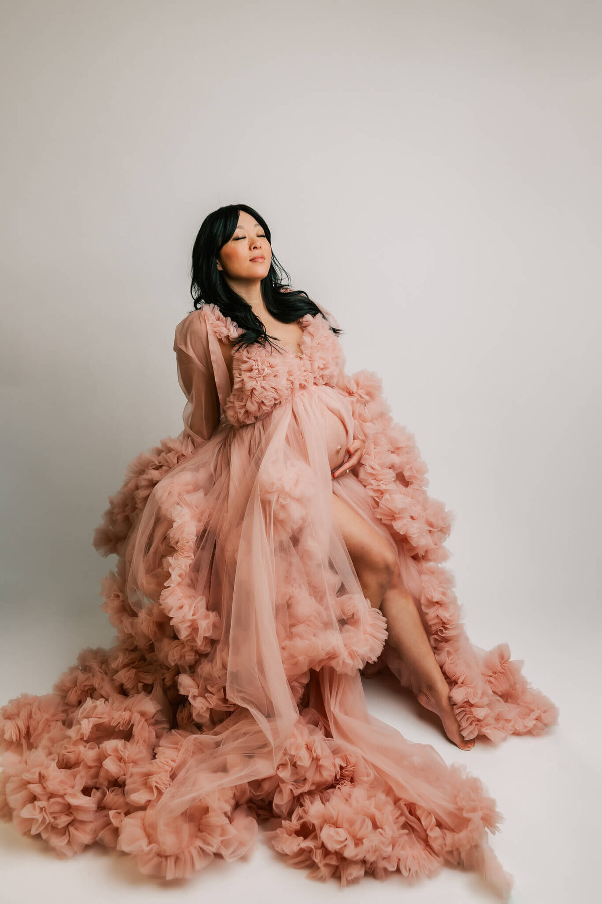 expecting mom posing in fine art studio maternity session in portland oregon wearing a pink tulle gown sitting on a stool with her leg pointed
