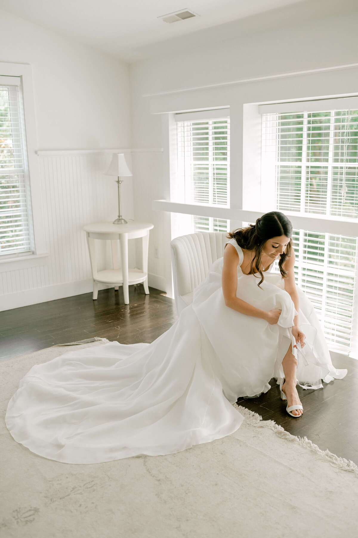 Bride in her bridal suite sitting in a chair to put on her shoes, taken by Rachael Mattio