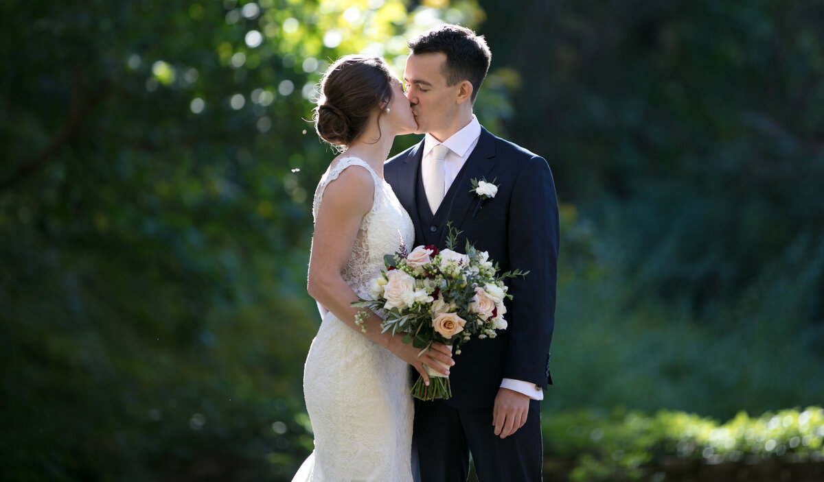 A bride and groom  sharing a kiss on their wedding day at the Avon Old Farms hotel in Connecticut. The bride, holding a bouquet of blush roses and assorted greenery, wears a white lace wedding dress with her hair styled in a lose updo. The groom is dressed in classic navy suit with a white boutonniere. They are set against a lush green backdrop with sunlight filtering through the trees., highlighting the romantic moment. =photograph by Hans Gonzalez