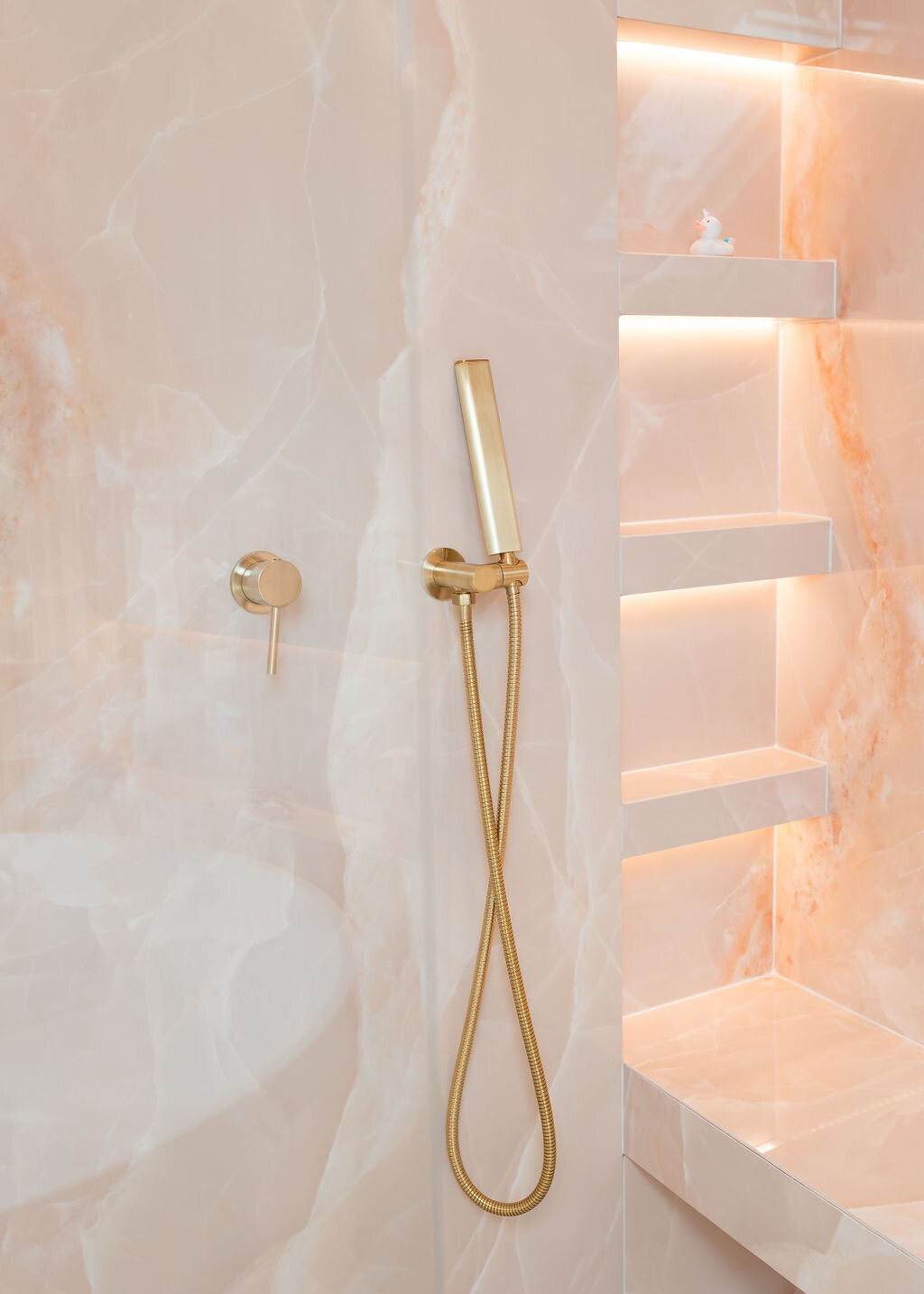 A luxurious glass-enclosed walk-in shower, featuring pink marble walls, gold hardware, built-in shelving, and a bench.