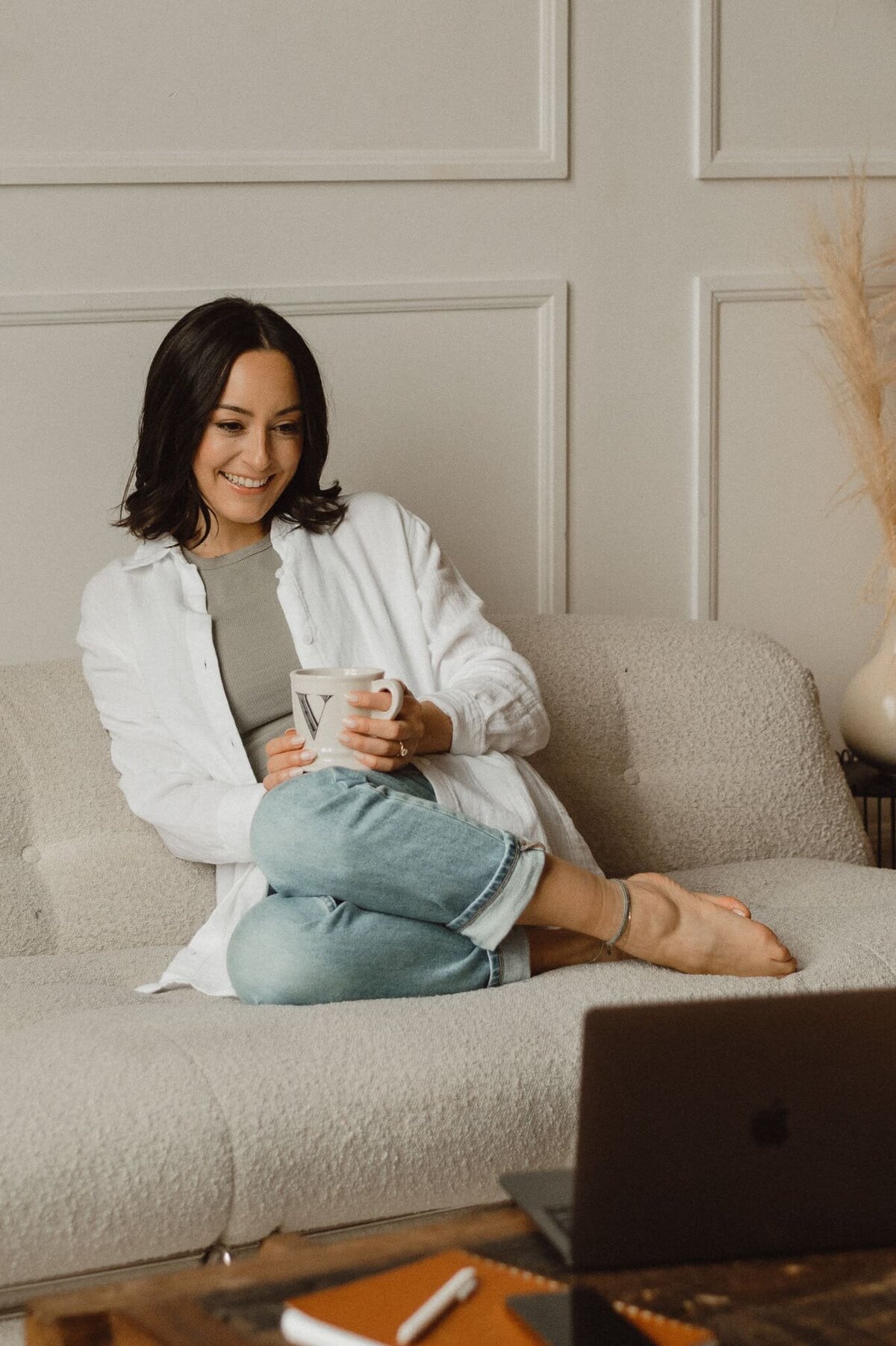 Vickie Chountalos of VERVE & COLOR holding a warm cup of tea, cozy on a sofa, smiling while looking at her laptop during a client meeting.