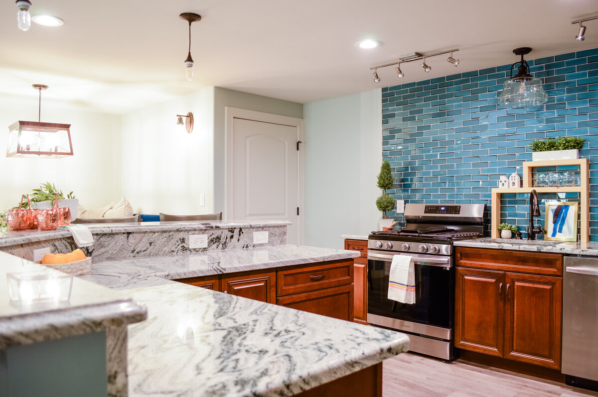 This image shows a kitchen with a mix of modern and rustic elements. The countertops are a light gray and white granite, contrasting with the warm brown wooden cabinets below. A kitchen island with a light blue base and the same granite countertop extends into the foreground. The backsplash behind the stove and sink is a vibrant teal subway tile, illuminated by track lighting. A stainless steel gas range with a white towel hanging from the handle stands against the teal backsplash. To the right of the stove, a stainless steel dishwasher is visible. A wooden shelf unit with greenery and clear glass jars sits on the countertop near the sink. Above the sink, a clear glass pendant light hangs. The walls are painted in a light gray, and the flooring appears to be light-colored wood-look planks. In the background, a dining area with a light-colored banquette and pillows is partially visible. Another pendant light with a dark frame hangs above the dining area. A white door is also visible in the background.