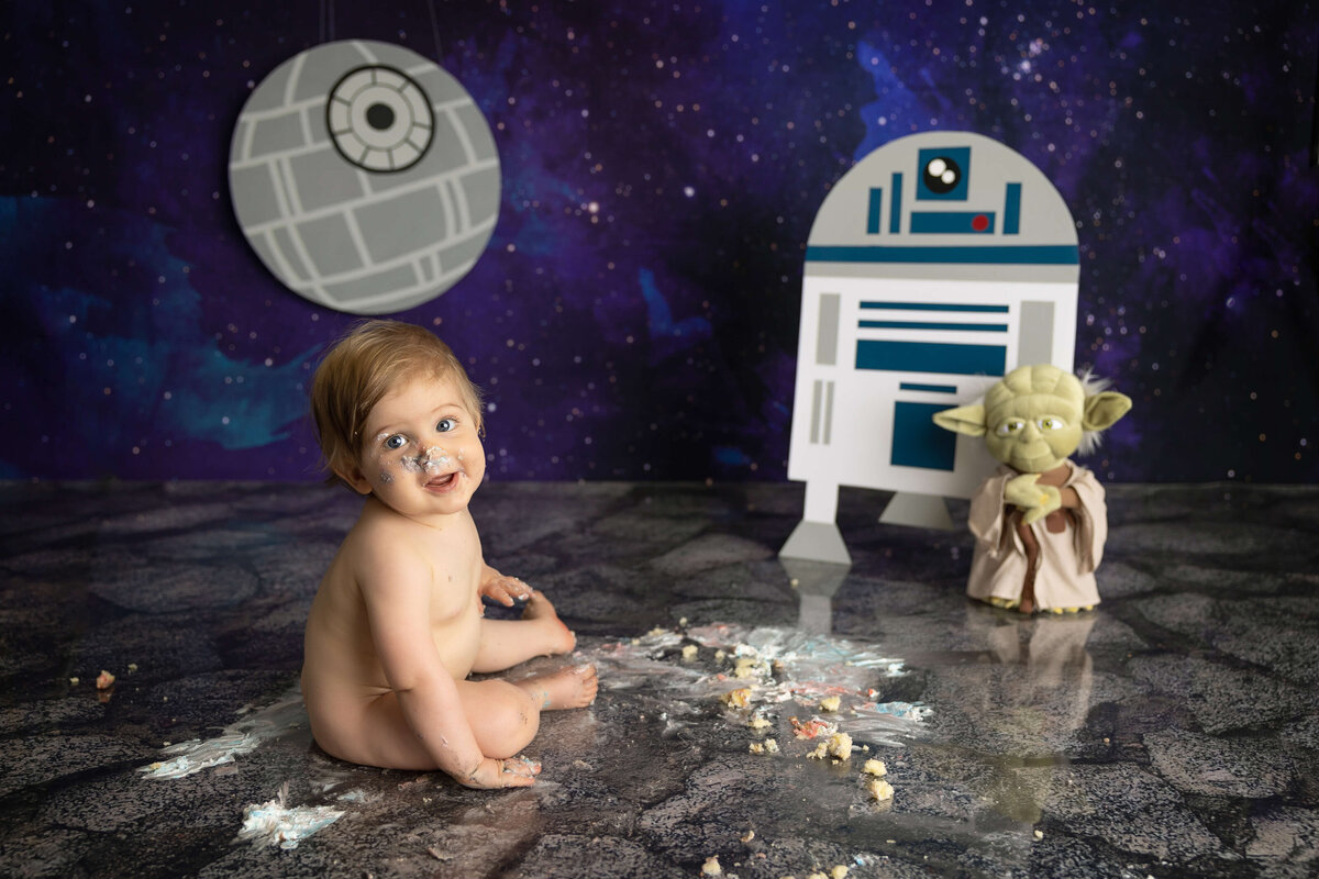 A baby smiling on a space-themed floor with cake smeared around sets the perfect scene for Jacksonville cake smash photography. Decor includes a paper R2-D2 and Yoda figure, alongside a Death Star wall decoration against a starry backdrop.
