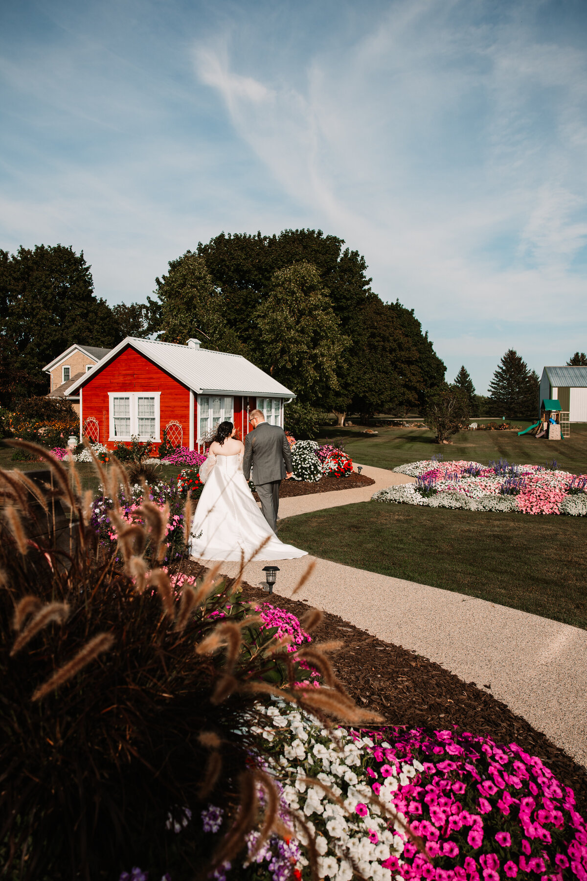 Newlyweds walk hand in hand on a garden sidewalk towards a red cottage