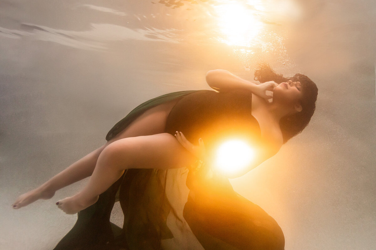 An enchanting underwater portrait featuring a woman in a dark green gown bathed in a radiant golden glow. The ethereal light highlights her serene expression, creating a breathtaking moment of beauty and calm.