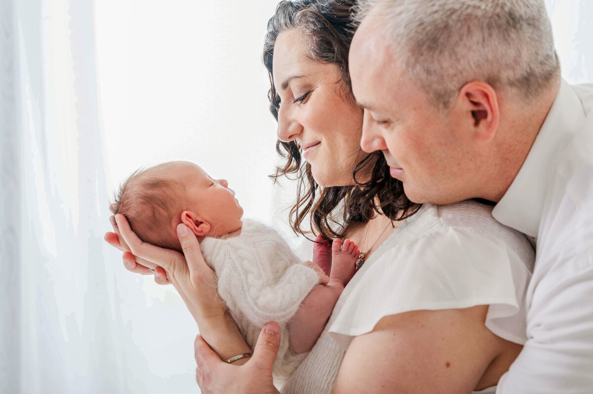 Dad is wearing a white dress shirt and looking over mom's shoulder at their newborn in mom's arms, while sitting in front of window with sheer curtains.