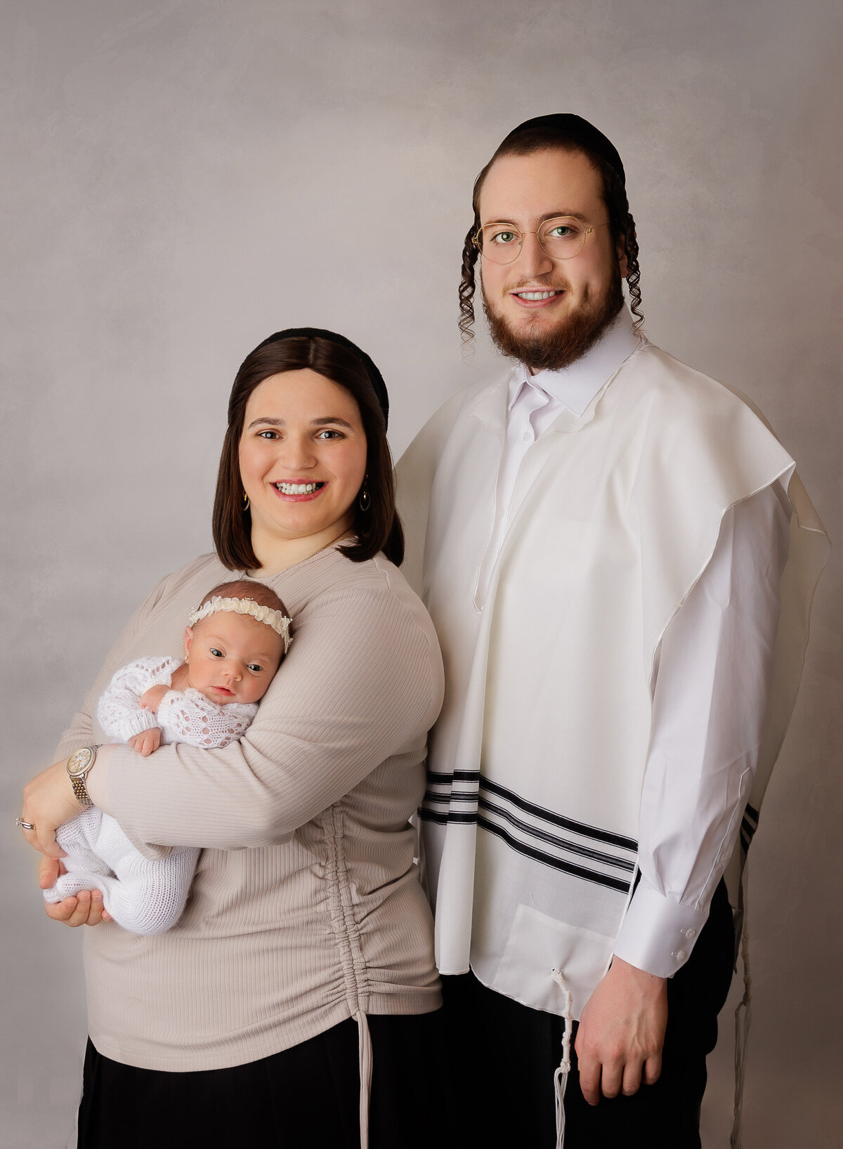 Mom and Dad pose for a newborn portrait with brooklyn newborn photographer. Mom is holding baby girl and baby is awake and looking at the camera. dad is standing behind mom wearing a Tallit katan. Captured by top brooklyn newborn photographer Chaya Bornstein.