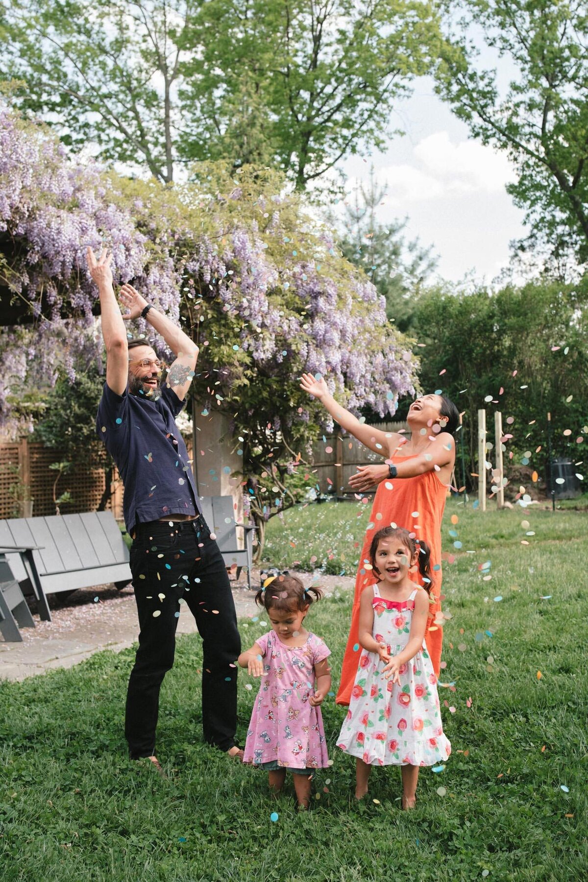 family throwing biodegradable water soluble rainbow confetti with purple wisteria in the background