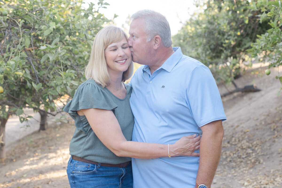 A man wearing a light blue shirt kissing his wife on her temple as she smiles with her eyes closed, her arm around his waist. Captured in lemon groves in Tustin.