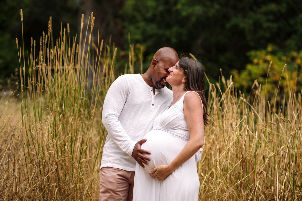 husband leaning into pregnant wife and touching her belly as she smiles in front of tall yellow grass