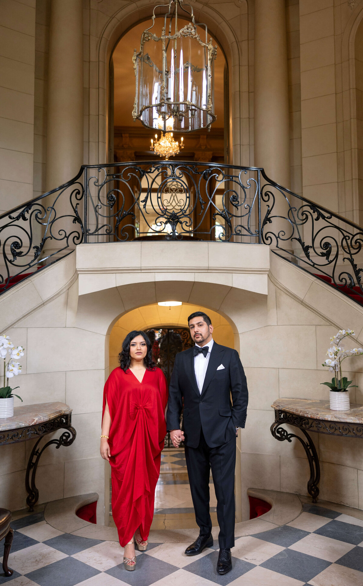 A couple stands hand in hand in a luxurious setting. The woman is wearing a red dress, and the man is in a black tuxedo. They are posing in front of a grand staircase with intricate railings and a large chandelier overhead.