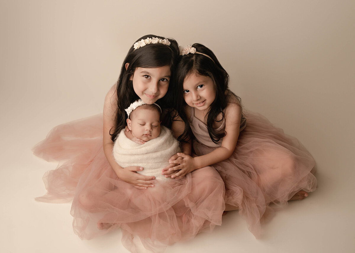 Two big sisters are holding their newborn baby sister for a photo captured at her newborn photoshoot. Big sisters have their heads resting together and smiling at the camera as the new baby is sleeping on the oldest sister's lap. Captured by best Murrieta newborn photographer Bonny Lynn Photography.