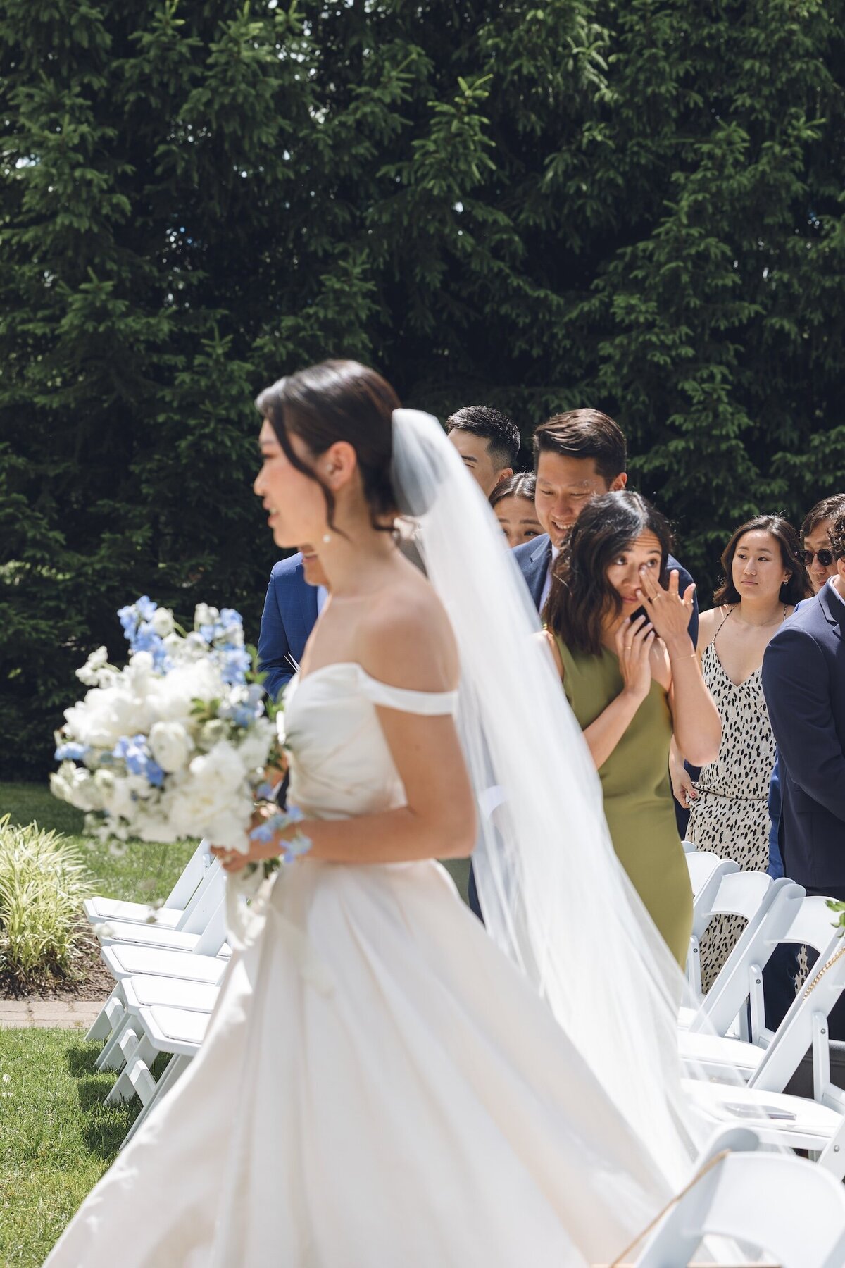 Bride walking down outdoor aisle holding white and blue bouquet