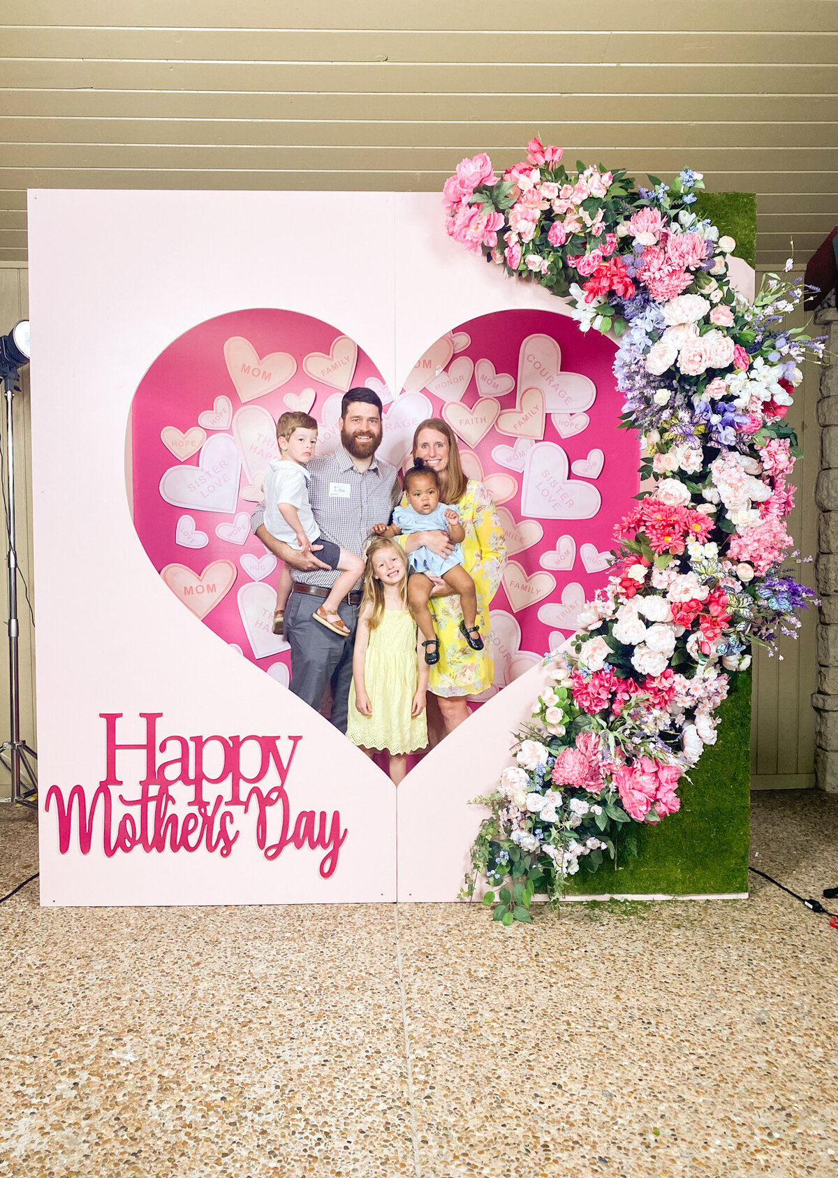 Family posing for a photo in a Happy Mother's Day pink heart shaped photo booth backdrop design with floral arch