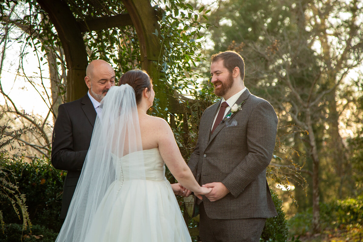 The bride and groom smiling while holding both hands right before they kiss at their wedding ceremony .