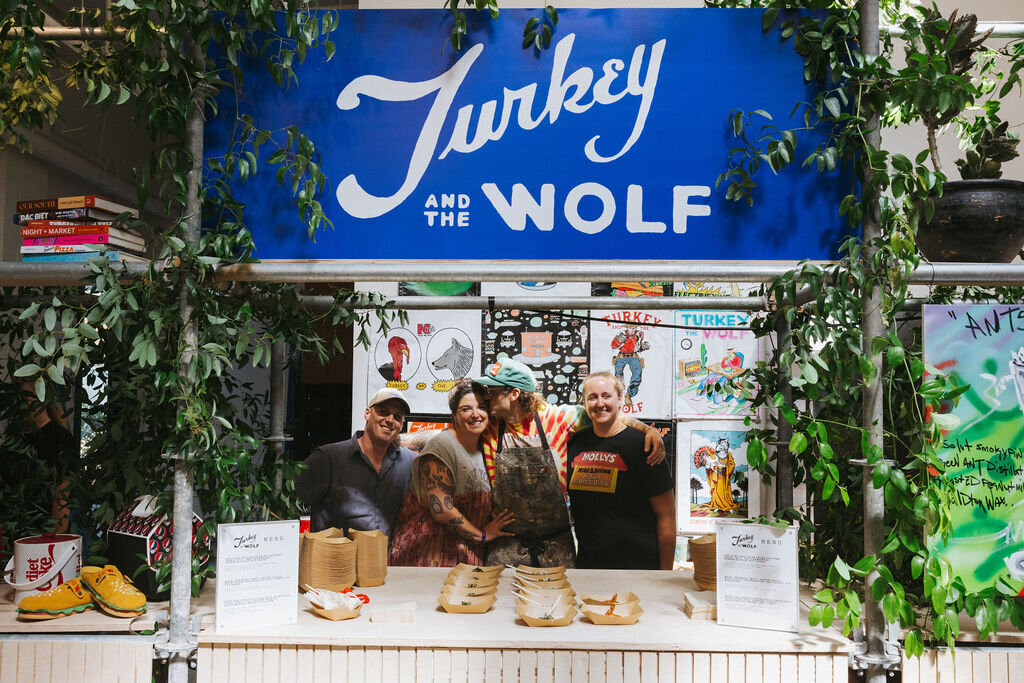 Turkey and the Wolf food stall with team members posing under a blue sign at the Absolut Vodka × Like Minded Creatures installation.