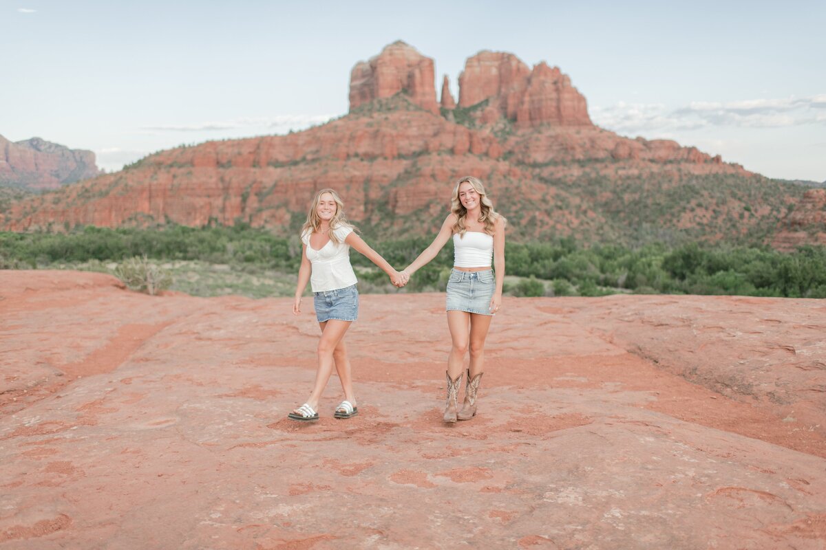 two senior girls holding hands walking on top of a rock formation in Sedona, AZ with cathedral rock in the background