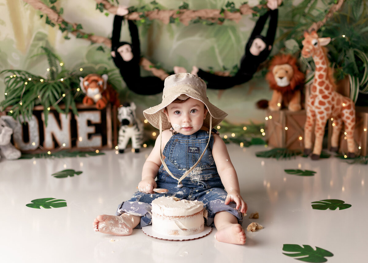 Safari jungle themed cake sash in West Palm Beach and Boca Raton, FL newborn and cake smash photographer studio. Baby boy is wearing overalls and canvas safari hat with cake on his overalls. He is looking at the camera. In the background, there is a jungle themed backdrop, leaves, and stuffed jungle animals.