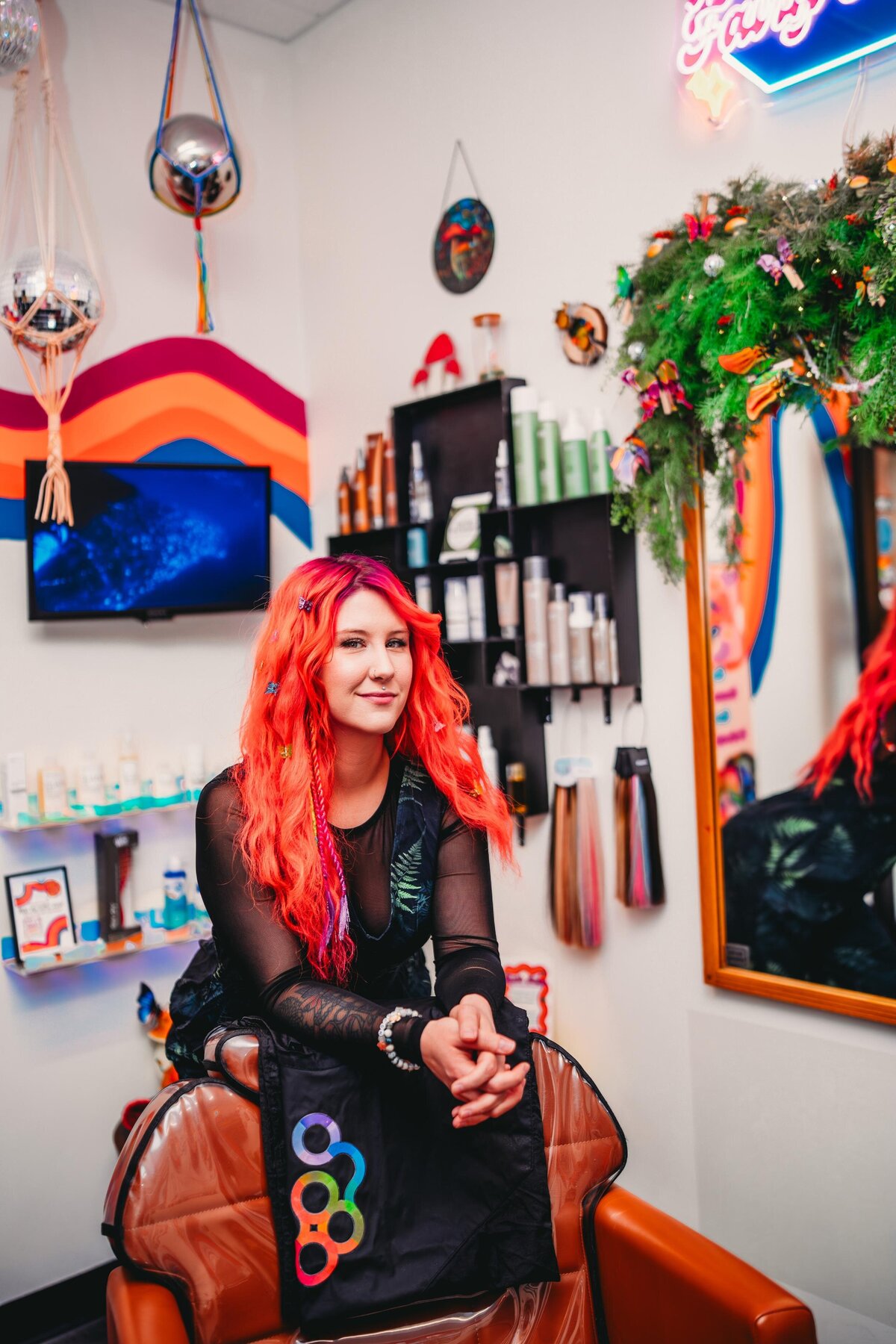 A moody portrait of a hairstylist with fiery red hair, sitting in a colorful salon with eclectic decor.