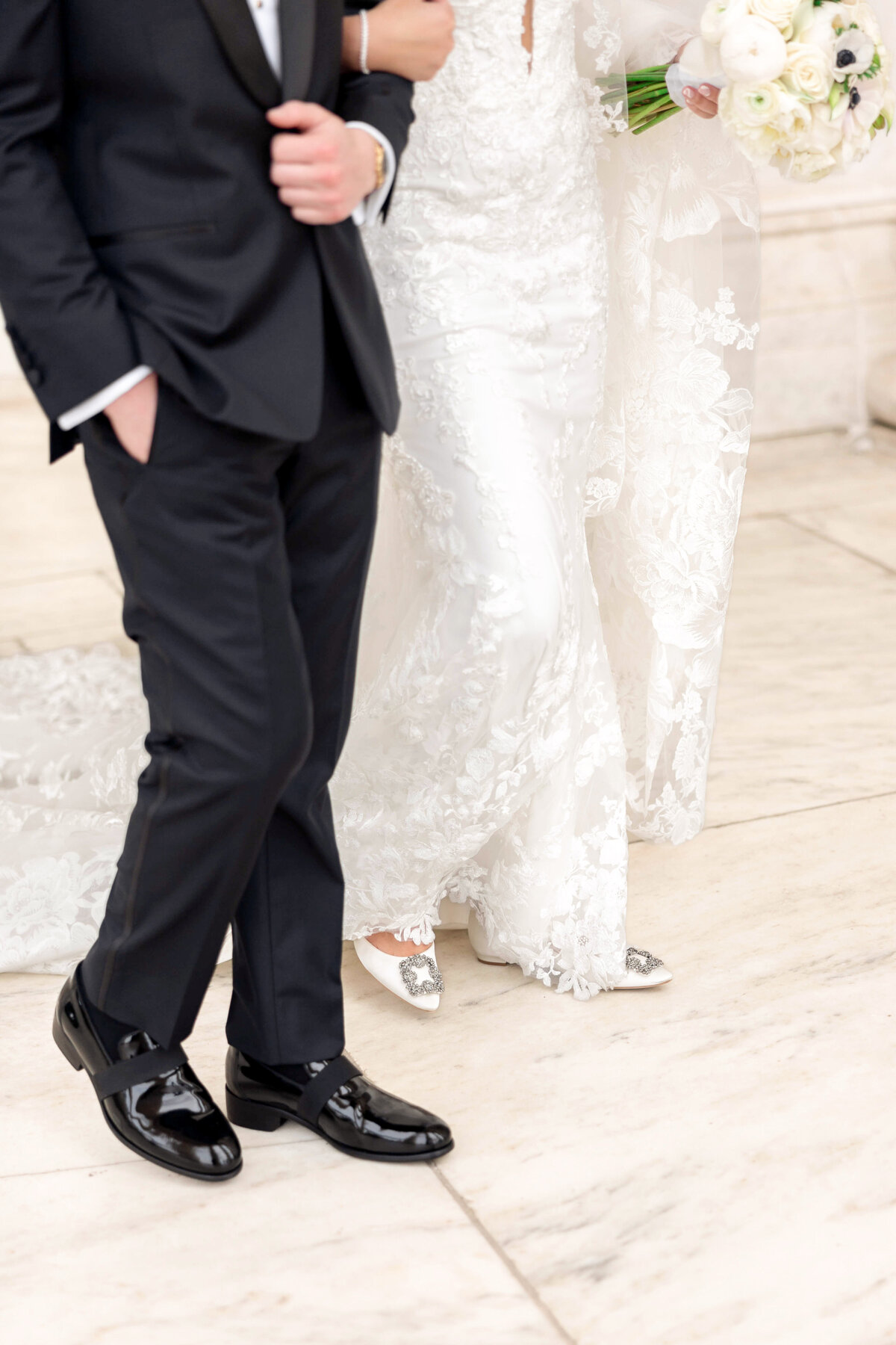A couple walking down a marble path. The groom is in a black suit and shiny shoes, while the bride wears a white lace wedding dress and holds a bouquet of white flowers. Their faces are not visible.