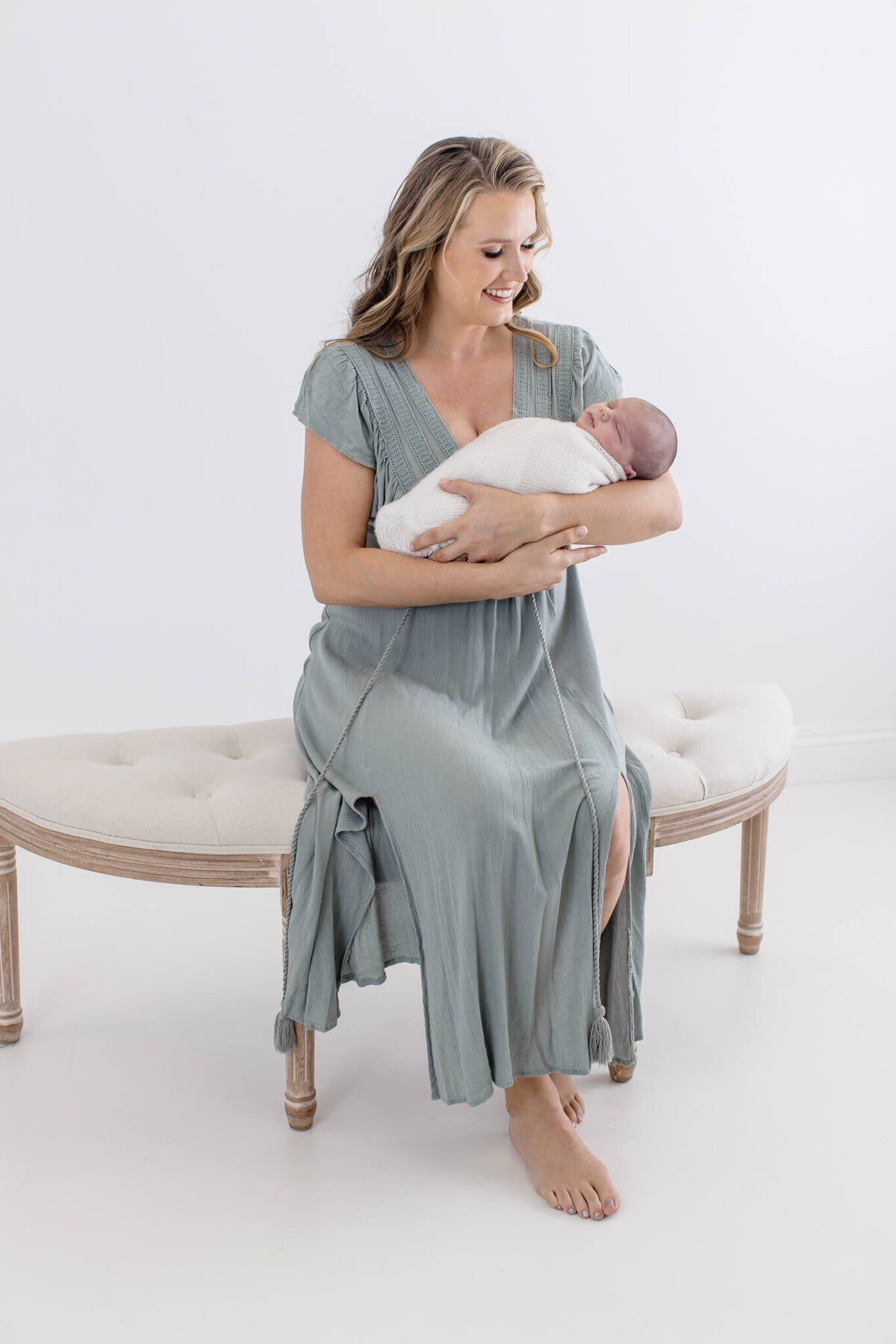 A woman in a light blue dress sits on a padded bench in a white room, holding a swaddled baby. She gazes down at the baby with a gentle smile. The setting is calm and serene.