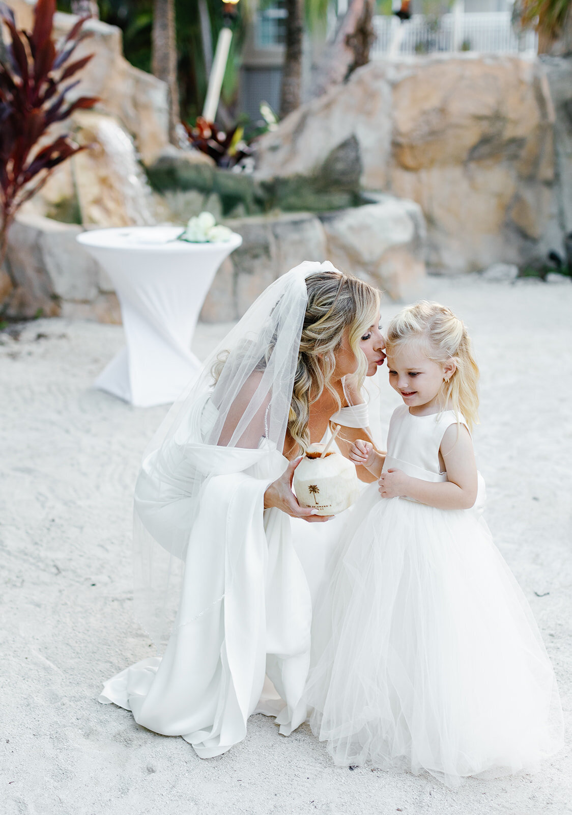 Monica kneels beside the flower girl, enjoying a refreshing coconut drink at their beachside wedding in Islamorada.