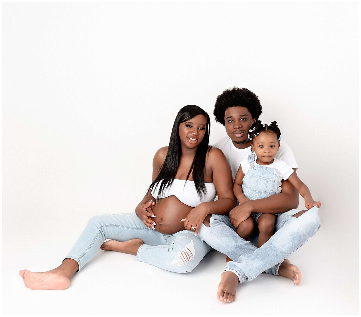 Family of three, including a pregnant woman, her partner, and their young girl, pose together in a professional studio setting on white backdrop for a maternity family session.