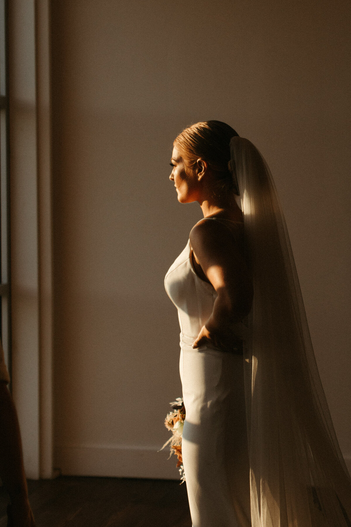 Modern bride in sheath dress and cathedral veil gazing out the window during sunset on her wedding day