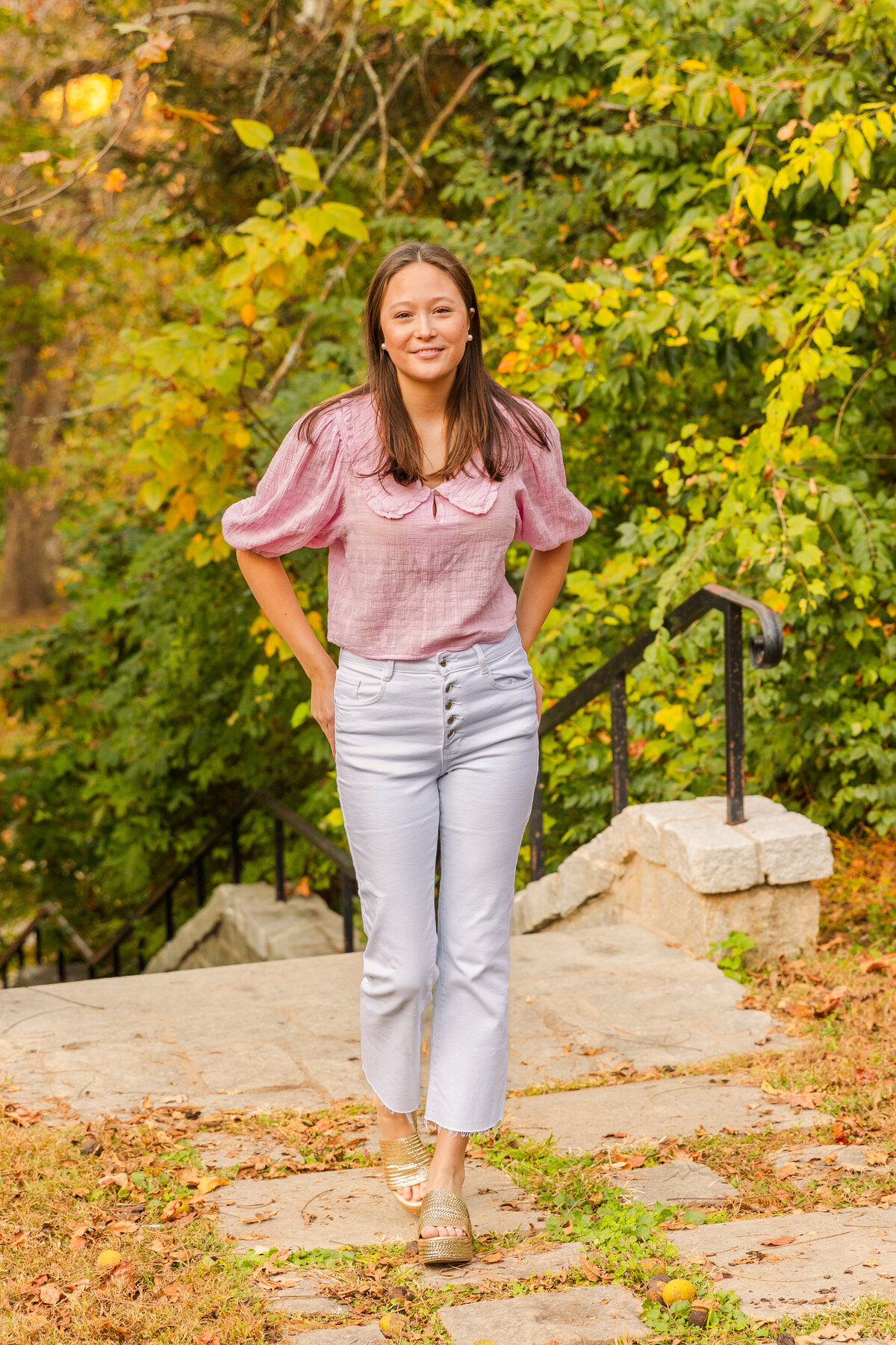 senior girl wearing white jeans and pink top walking towards the camera and hands on back pockets on park path