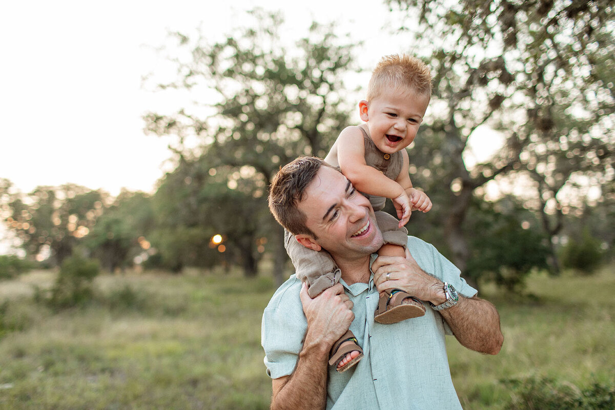 Outdoor candid portrait of a child laughing while sitting on their dad's shoulders, captured by San Antonio photographer Sara James.