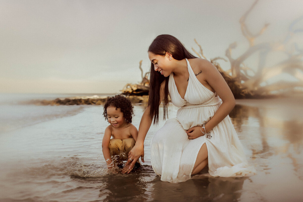 Woman in a white dress and a child play in shallow water at a beach. Driftwood is visible in the background.