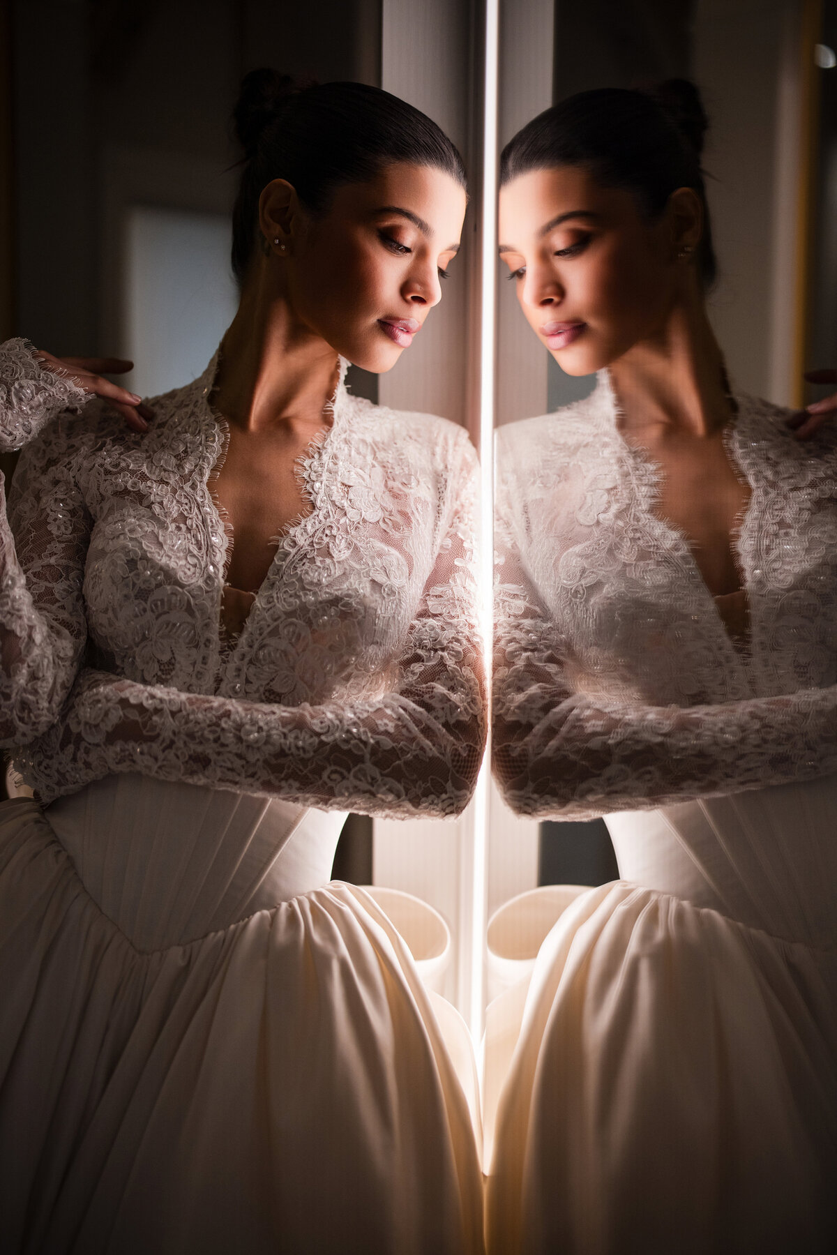 A model in a lace and satin wedding gown stands beside a mirror, her reflection softly illuminated by vertical light, wearing a corseted bodice and long lace sleeves. Photographed as part of the Pnina Tornai collection during New York Bridal Fashion Week, this image highlights the detail and elegance of couture bridal design.