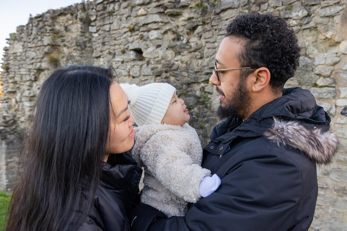 A couple holding and kissing their baby in front of a stone wall.