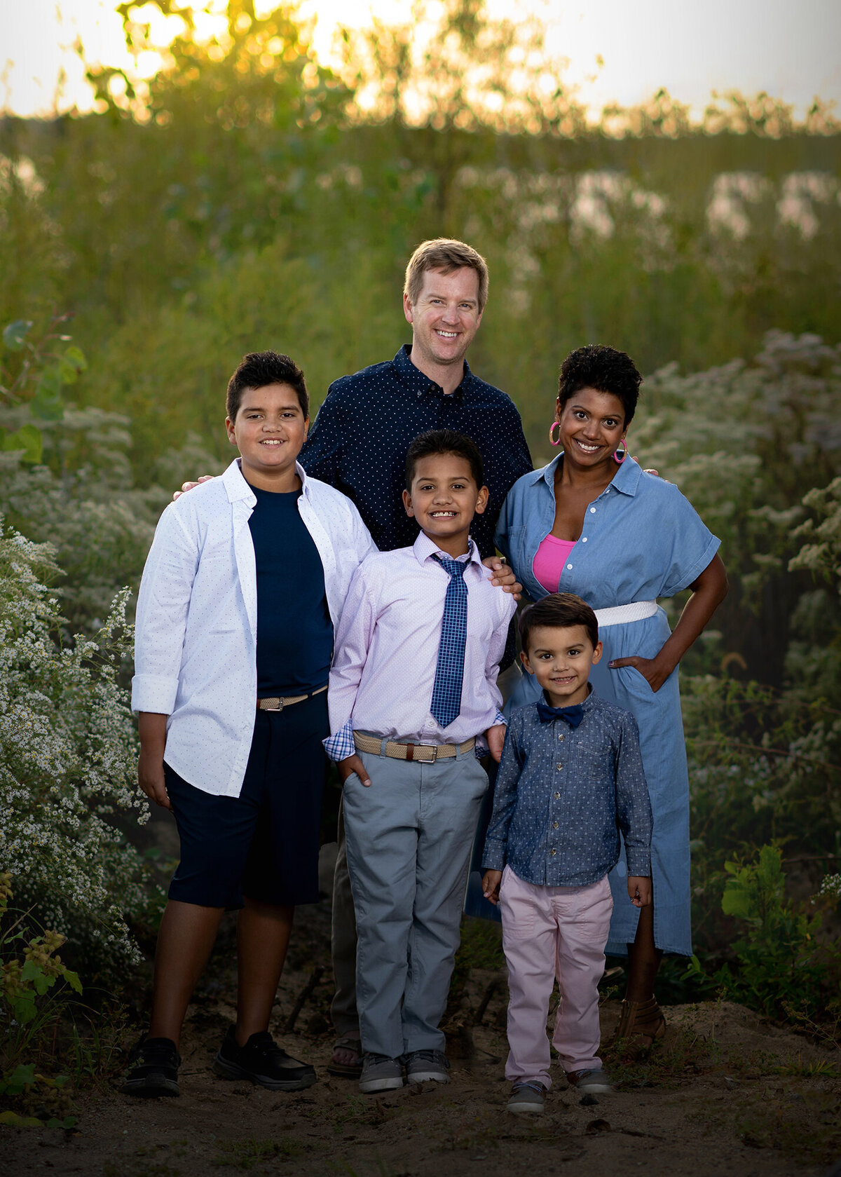 A family standing with green bushes around them puts their arms around each other and smiles for family photos