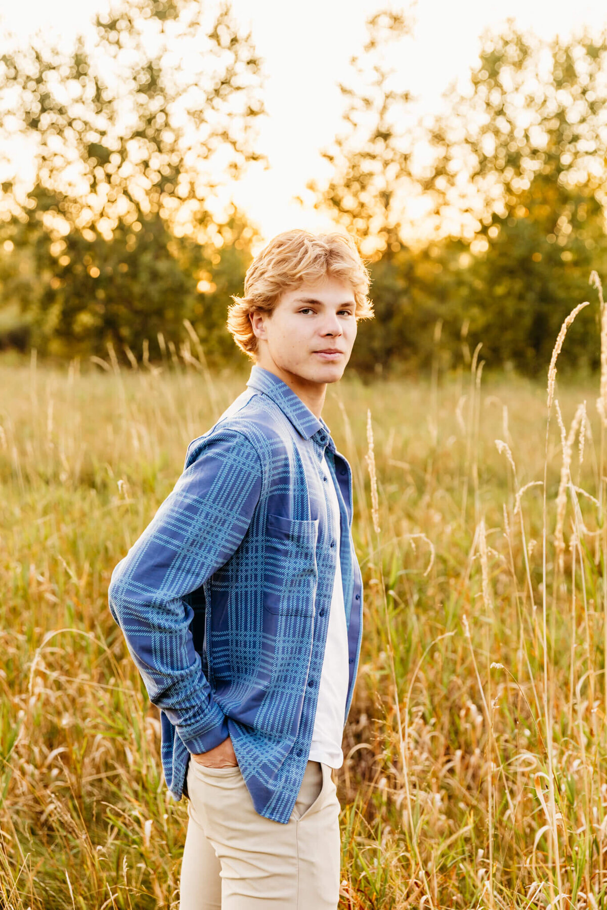 young man in a blue plaid shirt standing in a field at sunset and looking over .