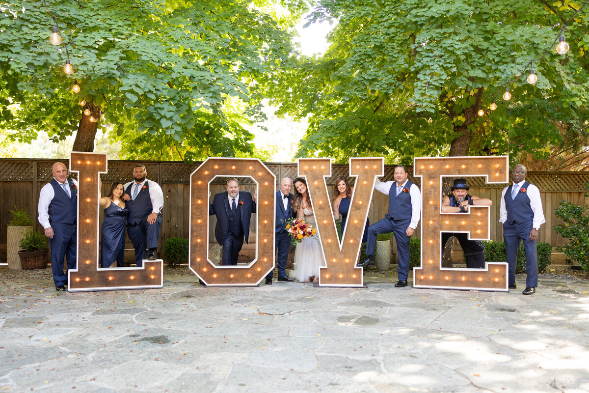 Bride and groom pose with groomsmen in the LOVE word sign at Forest House Lodge backyard, captured by philippe studio pro, a sacramento wedding photography studio.