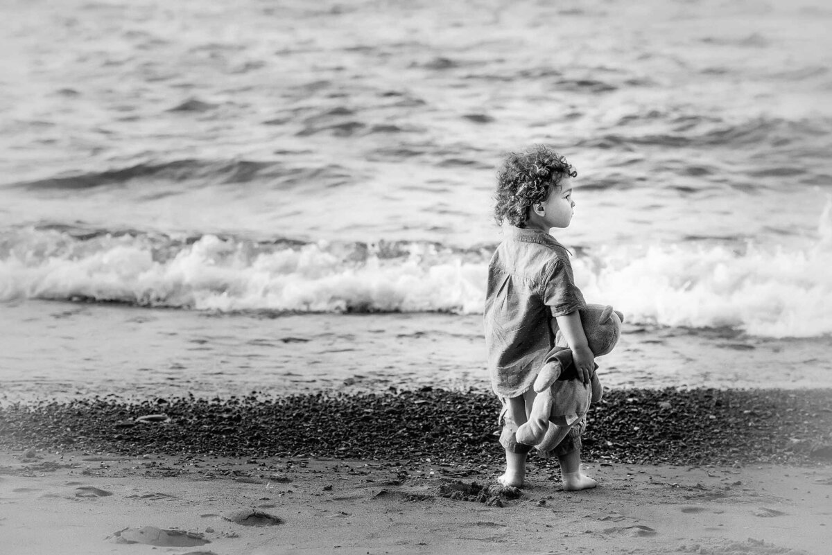 Toddler boy stands near the ocean looking into the distance holding his teddy bear.
