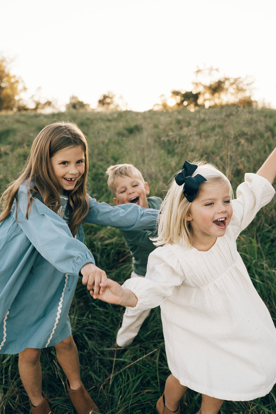 Three young siblings holding hands and spinning in a field during a documentary-style family photo session