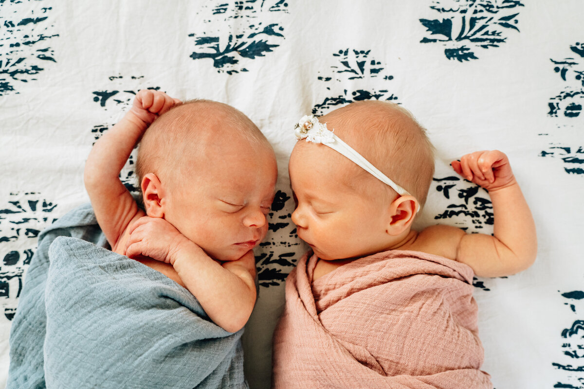 Two newborn twins swaddled in soft blankets, lying side by side on a patterned bedspread.