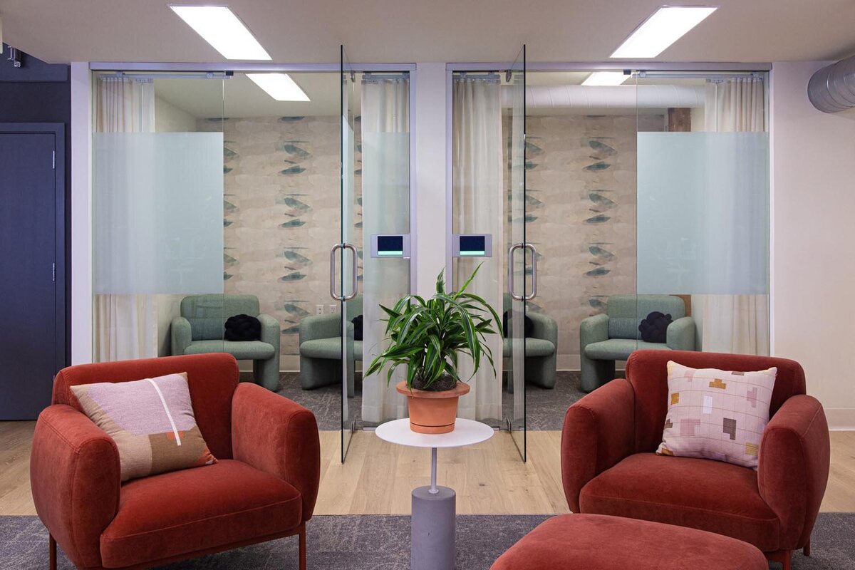 Seating area with rust-colored armchairs and glass meeting rooms featuring patterned wallpaper and frosted panels.