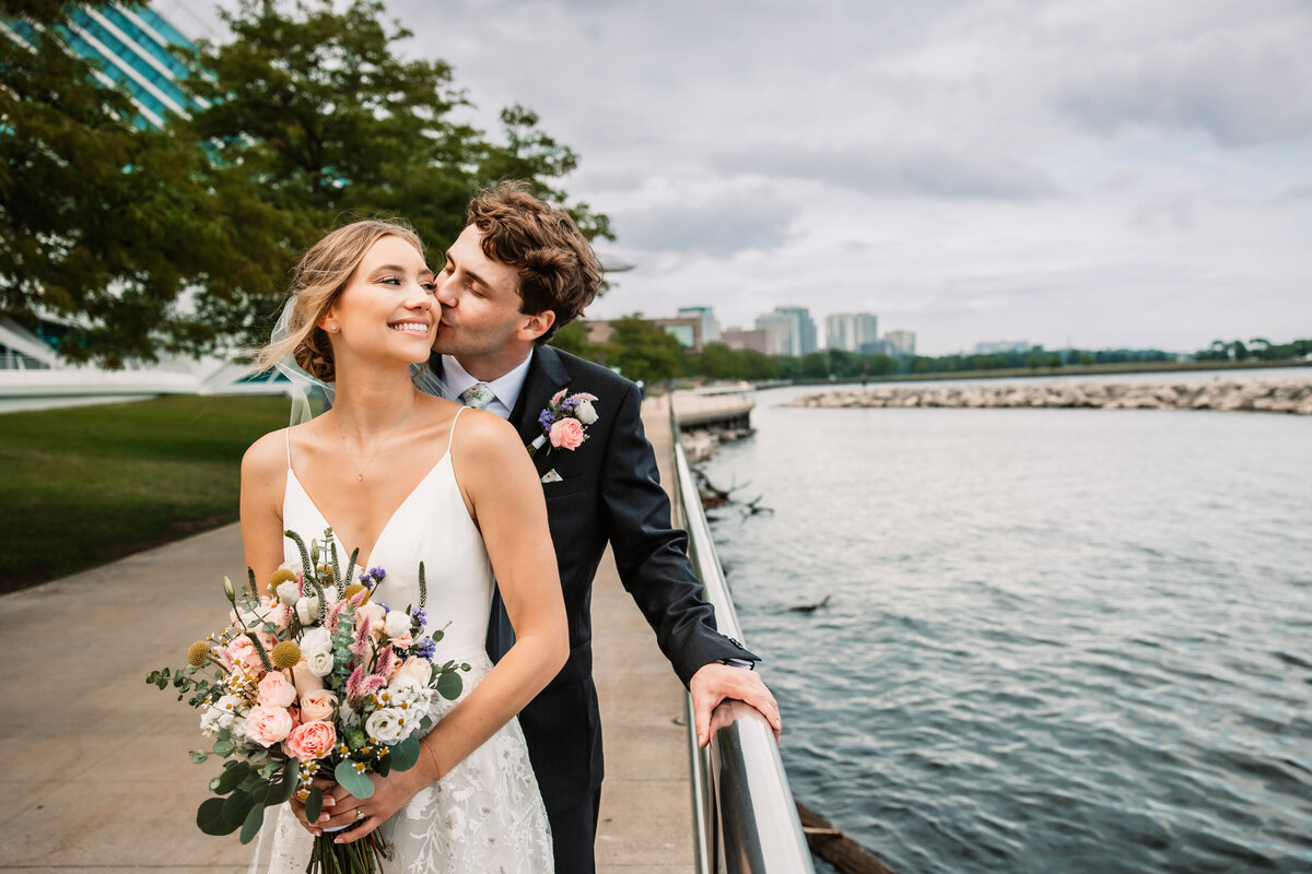 A groom kisses his smiling bride on the cheek from behind while leaning on a waterfront railing