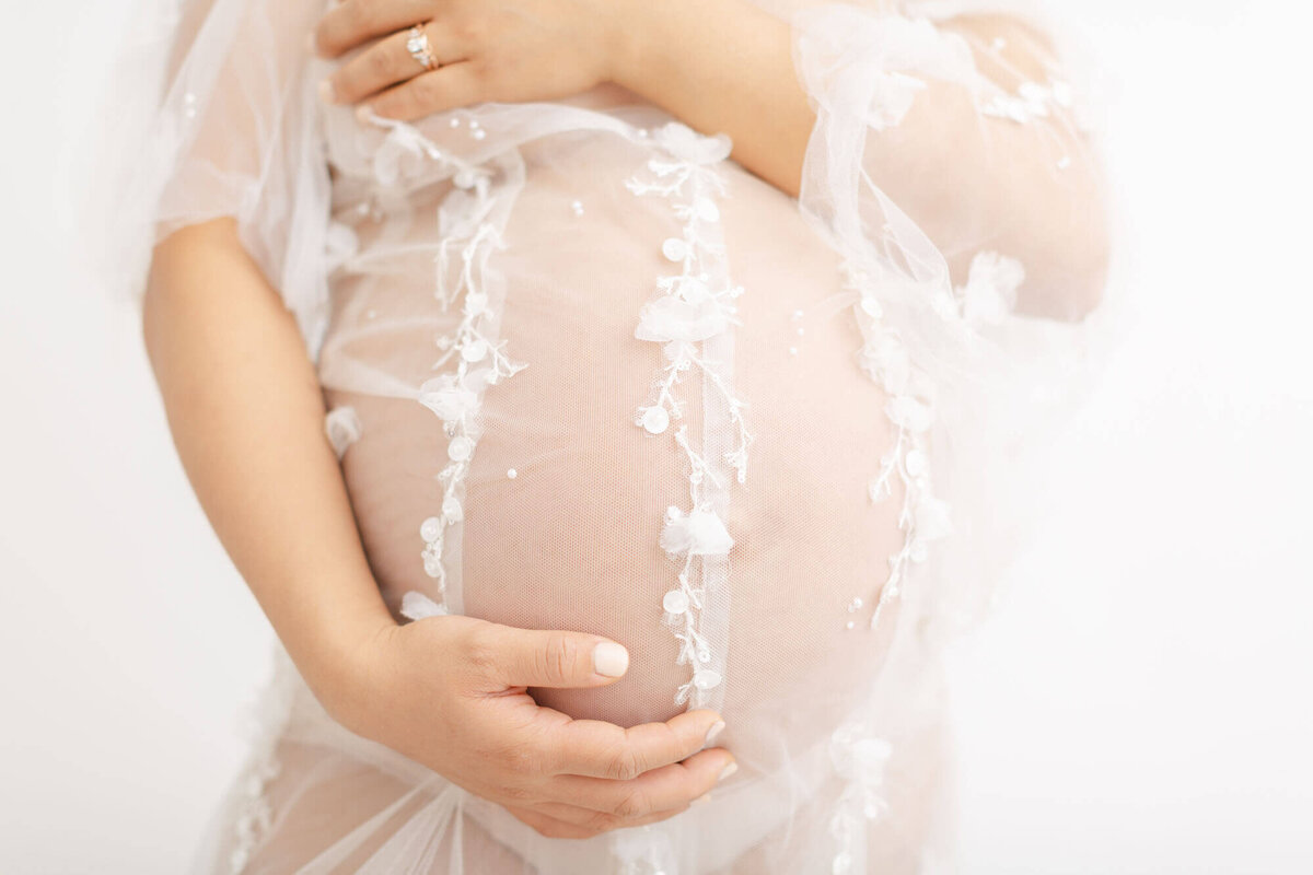Close up image of baby bump in a white photography portrait studio. Woman has one hand at the bottom of her bump and one hand is holding her chest. She is wearing a see-through tulle gown with hand-sewn bead work.
