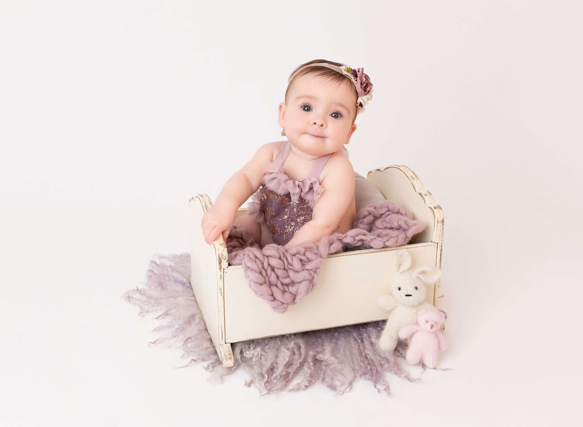 6-month-old baby girl in lavender outfit smiling at the camera during her NYC baby milestone photoshoot. Captured by a professional NYC baby photographer